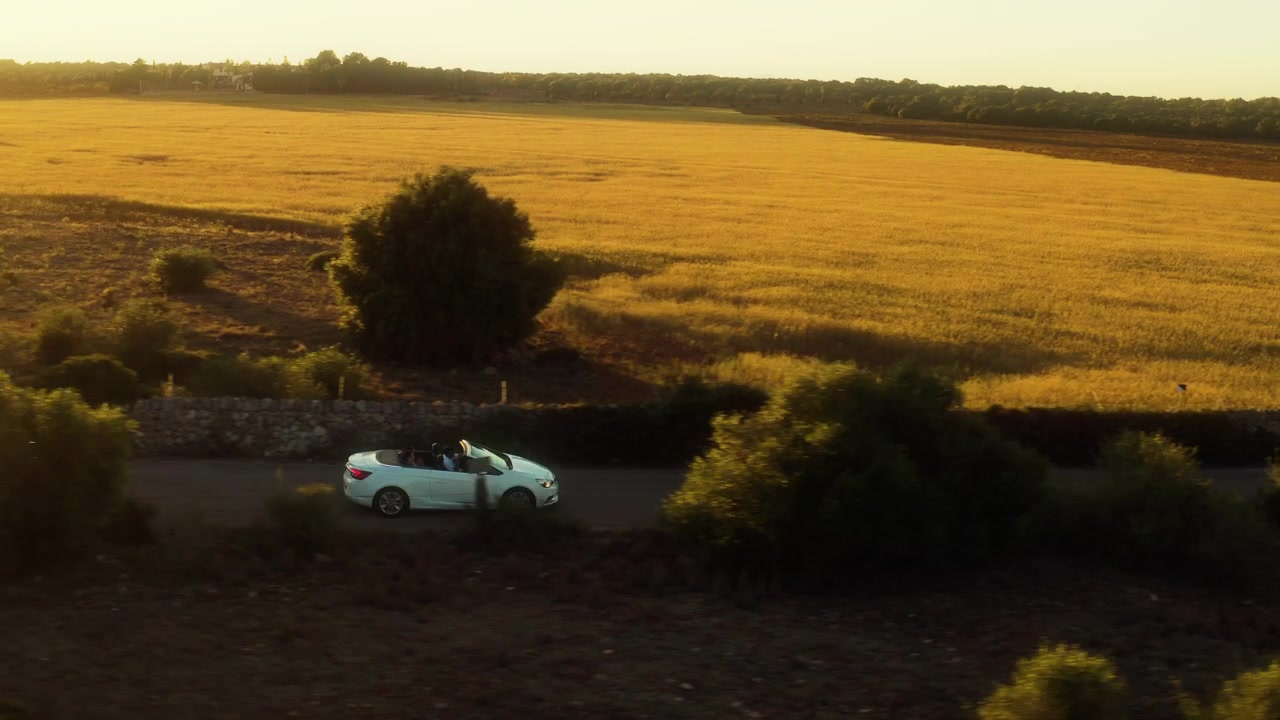 Una decappottabile bianca con due passeggeri passa accanto a un campo di grano dorato a Maiorca