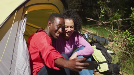 Smiling-diverse-couple-sitting-in-tent-and-taking-selfie-in-countryside