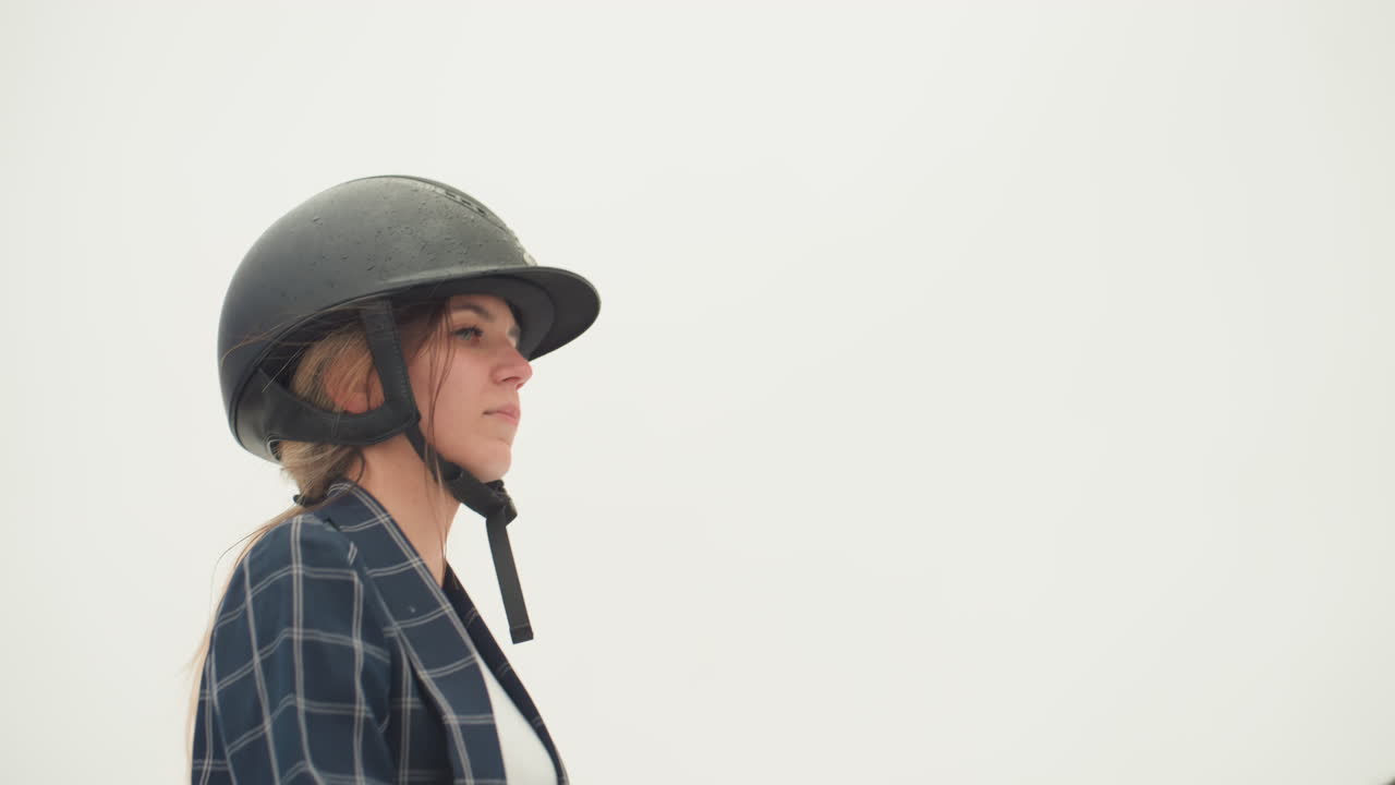 Seorang wanita fokus berlatih teknik berkuda dengan benar. Wanita Kaukasia tersebut menunggangi kuda dengan helm dan tali pengaman yang terpasang erat. Penunggang kuda wanita itu siap untuk instruksi, dengan tali kekang yang sudah diperiksa dan mengenakan pakaian pelindung.