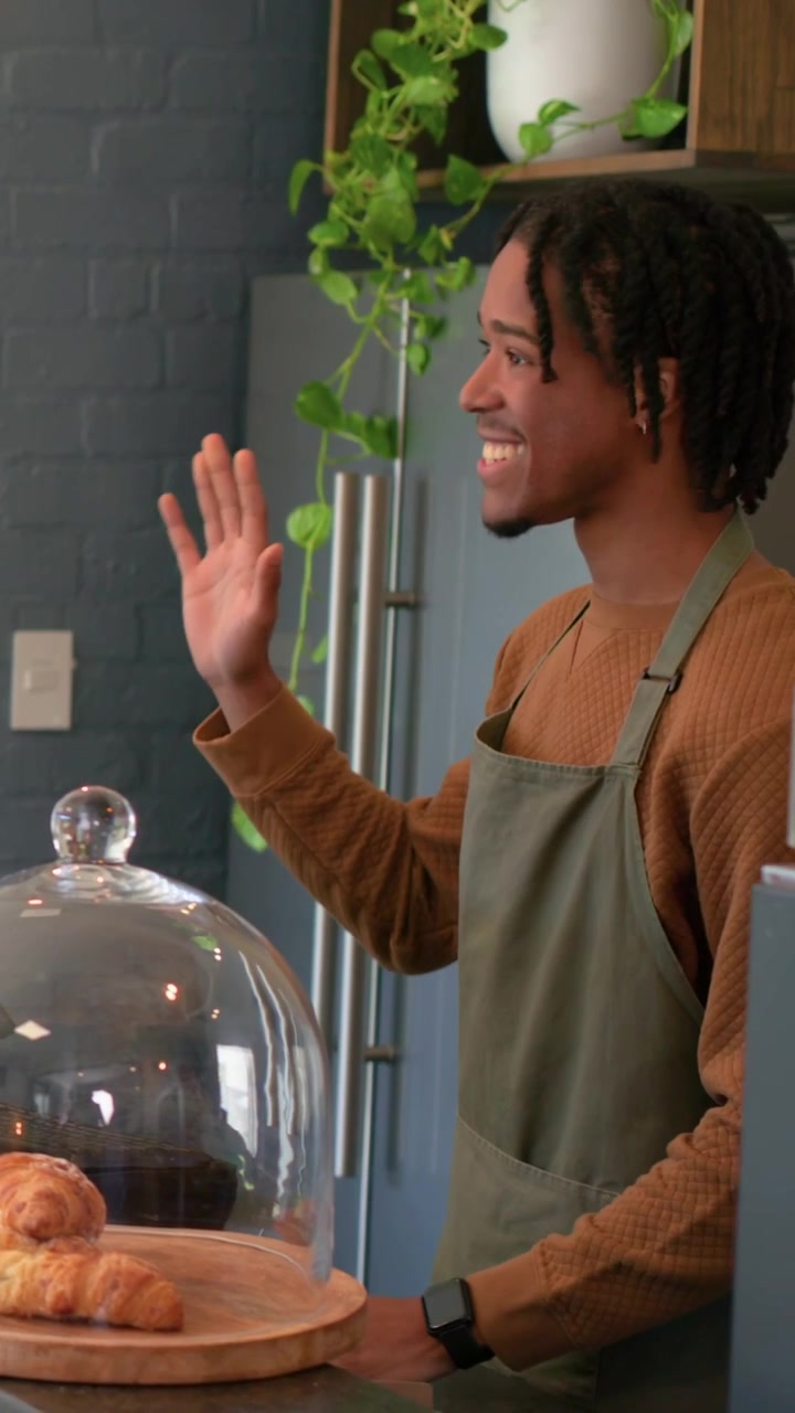 Video verticale: Uomo con grembiule verde e maglia marrone che serve un cliente in un caffè, elaborando una vendita con una cloche.