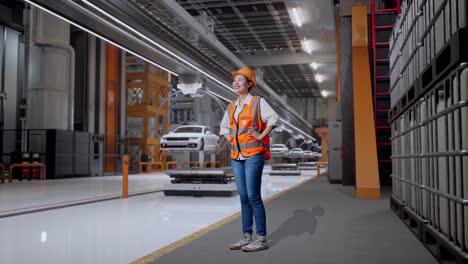 vista lateral completa del cuerpo de una ingeniera asiática que usa un casco de seguridad mirando a su alrededor mientras está de pie con los brazos akimbo en la fábrica de automóviles ev