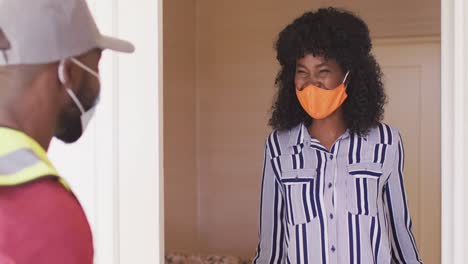 hombre de entrega con máscara de cara entregando un paquete a una mujer afroamericana con máscara de cara en casa