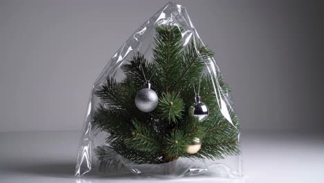 small christmas tree decorated with three ornaments, wrapped in transparent plastic film, resting on a white surface against a gray background, highlighting consumerism and holiday traditions
