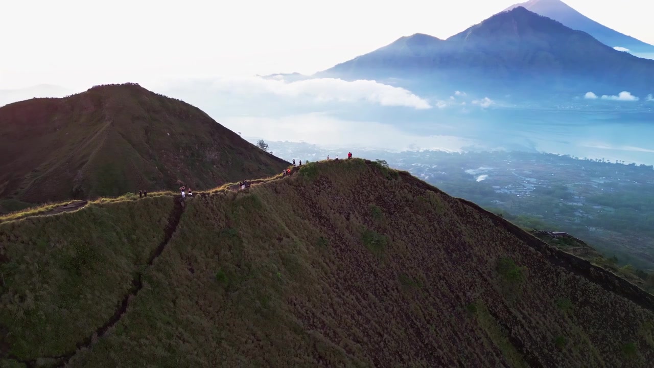 Riprese aeree con drone di escursionisti di montagna che esplorano una spettacolare cresta vulcanica sopra un lago craterico scintillante e una valle montana coperta di nebbia, nella luce brillante dell'alba.