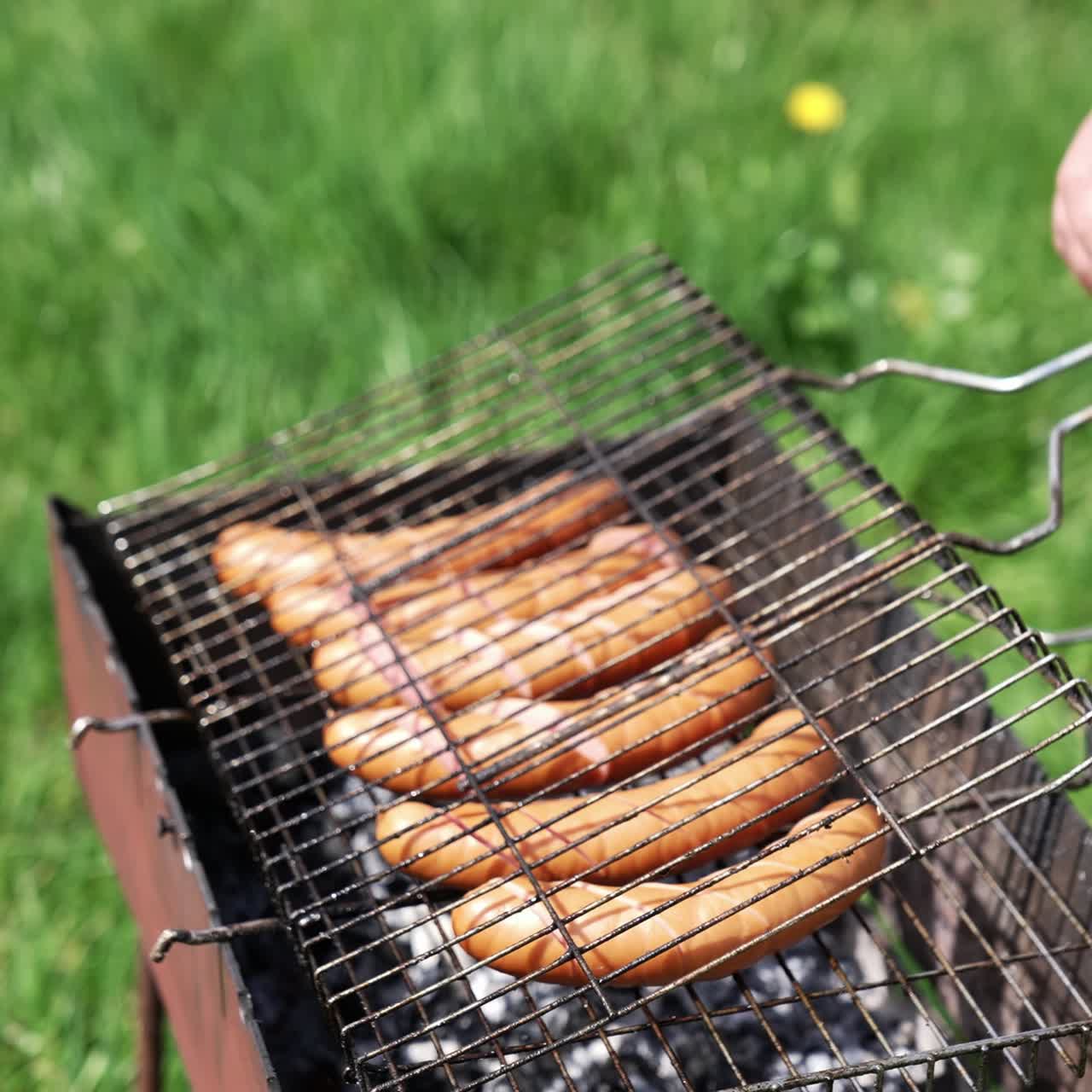 Man prepares juicy sausages in barbecue on picnic. Cook putting sausages into a grill for frying. Fried sausages are cooking on coals in the garden