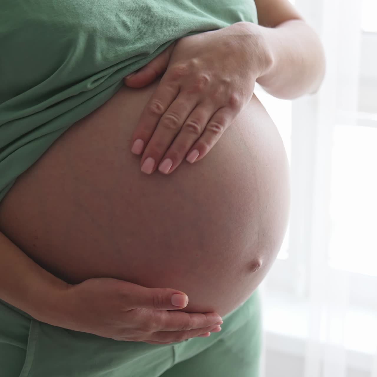 Futura madre de pie junto a la gran ventana. Sostiene su gran barriga con una mano y la acaricia con la otra. Primer plano.