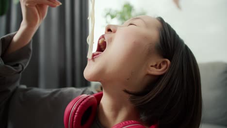 young asian woman in headphones eats noodles passionately with wood sticks at home, close up