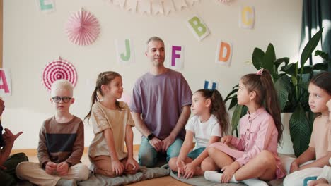 Unterrichtsstunde-Unter-Grundschulkindern-Mit-Dem-Lehrer.-Kinder-Im-Schulalter-Sitzen-Um-Ihren-Erwachsenen-Männlichen-Lehrer-Herum-Und-Hören-Ihm-Vor-Dem-Hintergrund-Der-Dekorationen-Zu,-Die-Dem-Ersten-Schultag-Gewidmet-Sind