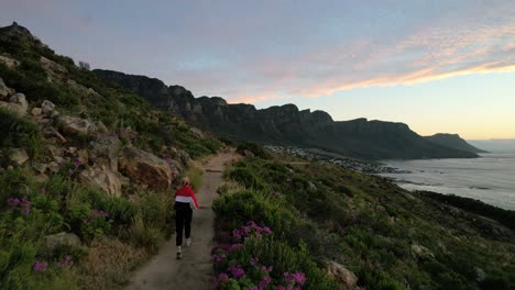 young blonde girl in red jacket running lions head trail at sunset towards 12 apostles in cape town, aerial