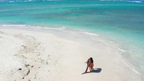 drone flies forward over woman tourist in bikini and sarong on tropical white sand beach, turquoise waters