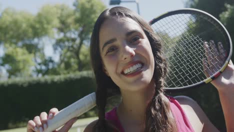 retrato de una feliz mujer biracial con una raqueta de tenis en el jardín en un día soleado