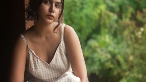 Young-woman-in-a-checkered-dress-gazes-contemplatively-out-of-a-window,-with-lush-greenery-in-soft-focus-behind