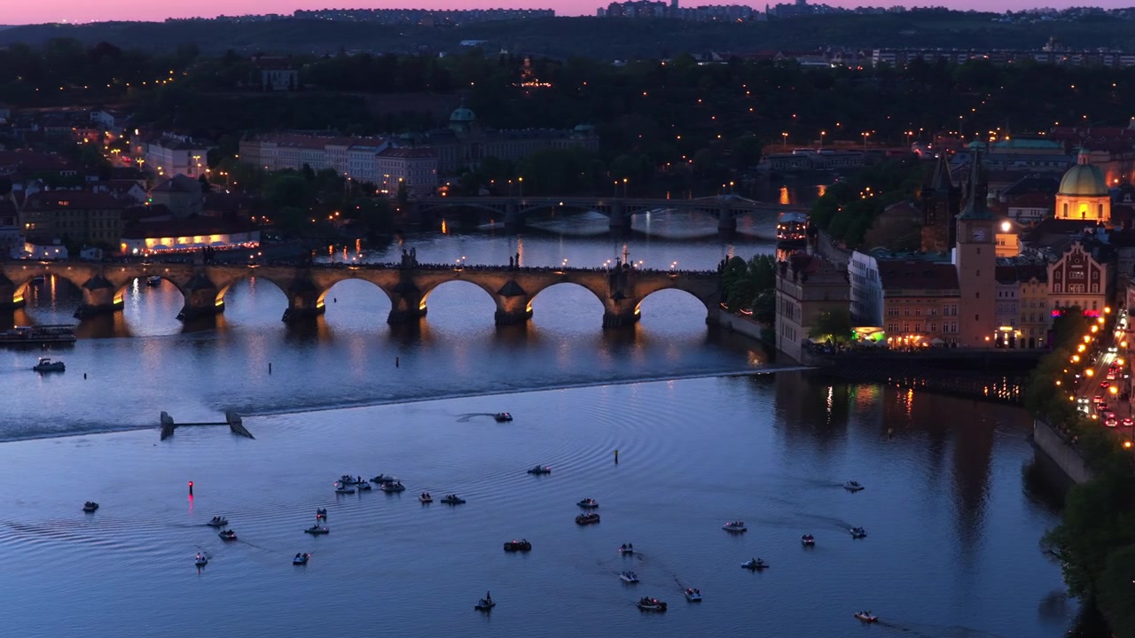 Vista aerea con drone di piccole imbarcazioni sul fiume Moldava al crepuscolo, con il Ponte Carlo e gli edifici illuminati del centro storico lungo il lungofiume, Praga, Repubblica Ceca.
