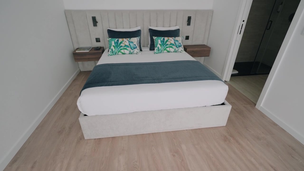 Camera da letto moderna e pulita con letto matrimoniale bianco, cuscini decorativi color verde acqua e comodini in legno.