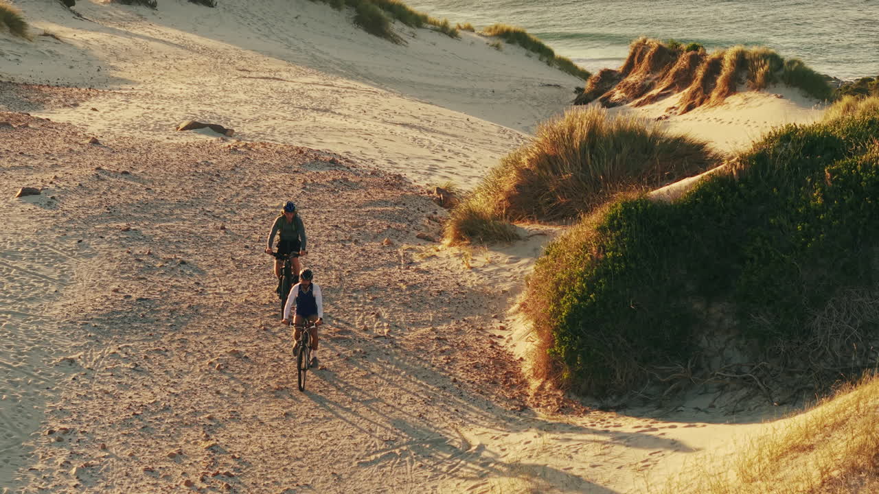 Due uomini in bicicletta sulle dune sabbiose in riva all'oceano al tramonto.