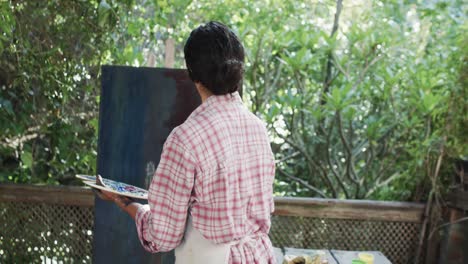 Artista-Birracial-En-Delantal-Pintando-Sobre-Lienzo-En-Un-Jardín-Soleado,-Cámara-Lenta