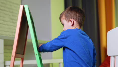 dark-haired toddler boy in blue sweater cleans the portable blackboard. side view portrait of the kid drawing at home.