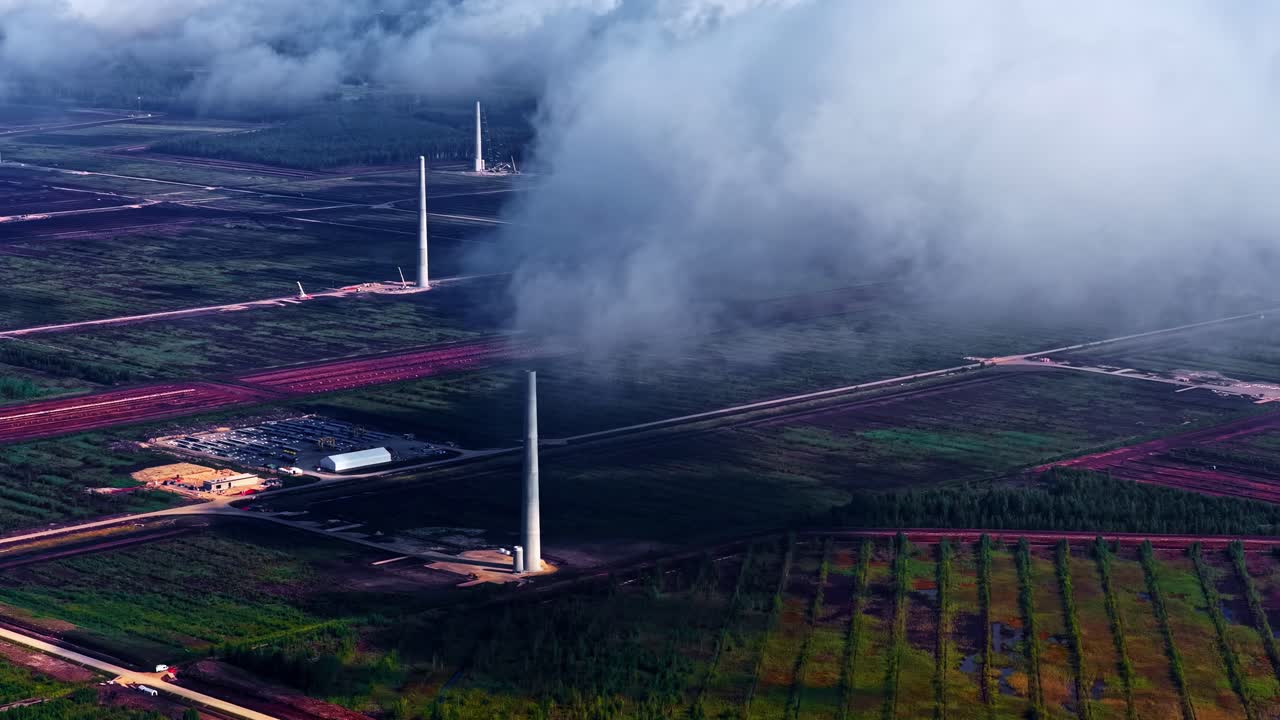Drone shot of renewable energy project site showing several tall white turbine towers standing in fields and forests of Latvia