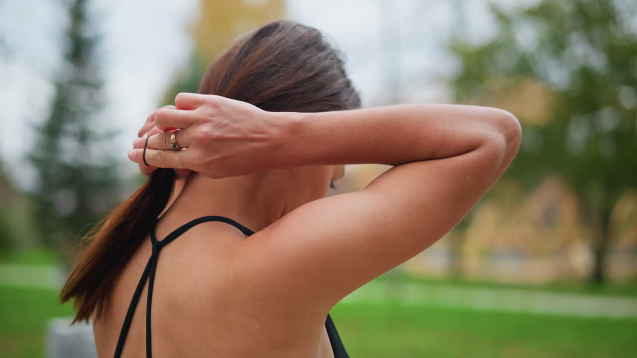 Back view of woman in black singlet packing her hair and tying it up, ready for workout or exercise, outdoors with green background and iron fence in view