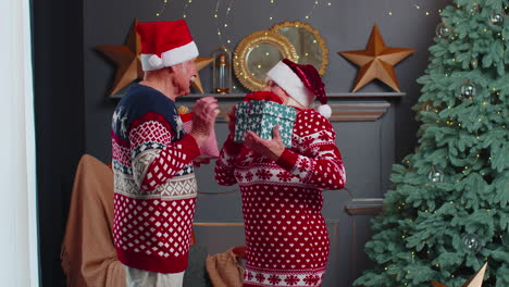 romantic senior couple presenting gift boxes to each other at home near decorating christmas tree