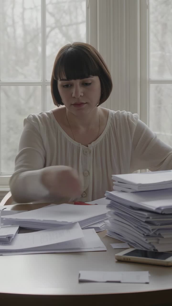 Vertical video: Seeing papers woman in top stacking them, sorting at table by window with red pen