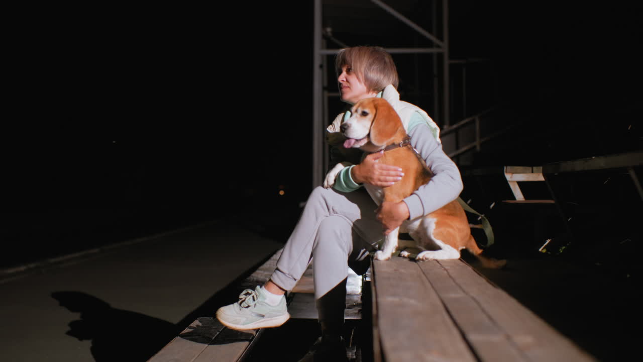 Sport woman dressed in sportswear seated on wooden bench holding her athletic beagle dog closely under stadium lights at night showing deep companionship and peaceful bond after training session