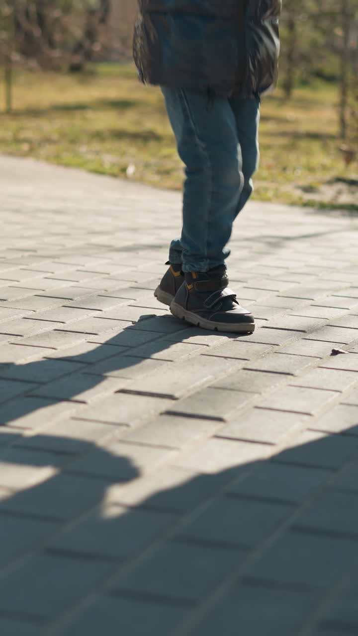 un primer plano de dos personas, un adulto y un niño, moviendo juguetonamente sus piernas en un camino pavimentado, ambos llevan vaqueros azules y botas, sus sombras se extienden a través del pavimento