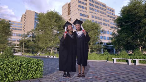 full body of asian woman students graduate in caps and gowns hugging each other, smiling, and showing diplomas in their hands to camera in front of a magnificent university building