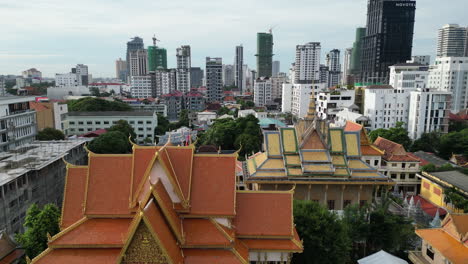 aerial video of phnom penh highlights vibrant orange temple rooftops, flanked by golden and green tones, set against dense city blocks and modern high-rise towers