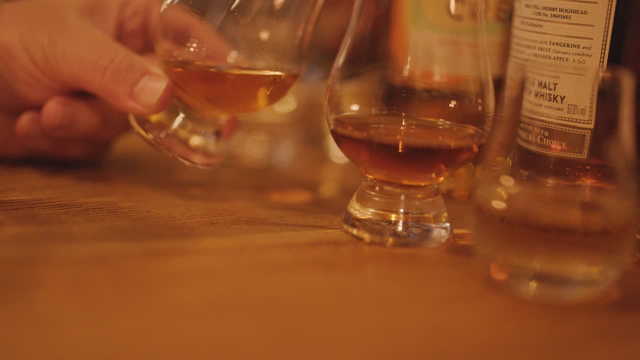 Hand swirling whisky glass on wooden table, warm lighting, surrounded by bottles and glasses