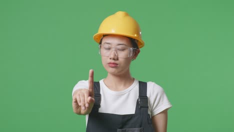 close up of asian woman worker wearing goggles and safety helmet disapproving with no index finger sign while standing in the green screen background studio