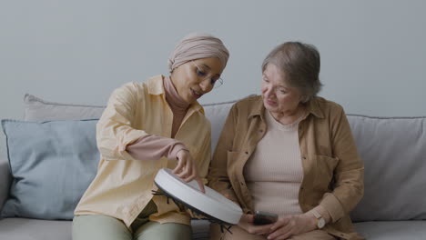 middle aged arabic woman explaining to a senior woman how to use the robot vacuum cleaner while sitting together on couch at home