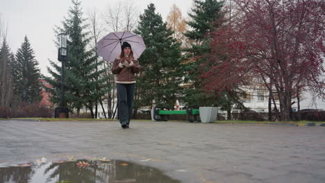 female walking peacefully on cold overcast day through paved park holding closed umbrella, dressed in black knit cap, brown shearling jacket, and black trousers, gentle snowfall