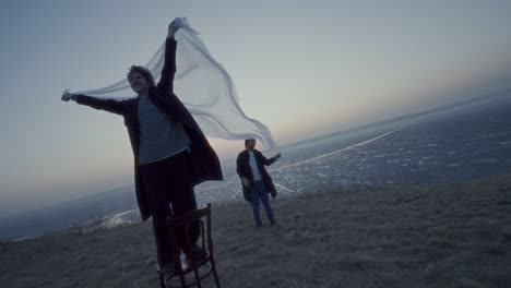 carefree woman standing on chair, holding fabric with boyfriend by frozen lake