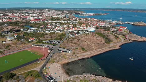 Hermoso-Paisaje-De-Casas-Y-Calles-En-El-Puerto-De-Pinnevik,-Lysekil,-Suecia-Con-Barcos-Navegando-Suavemente-Sobre-El-Agua-Del-Mar-En-Un-Clima-Brillante---Toma-Aérea