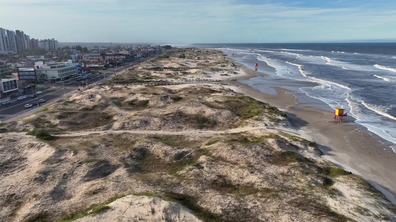 Sanddyner vid Praia Grande-stranden med en trägångväg och havskustlinje, samt flygfoto med framåtrörelse.