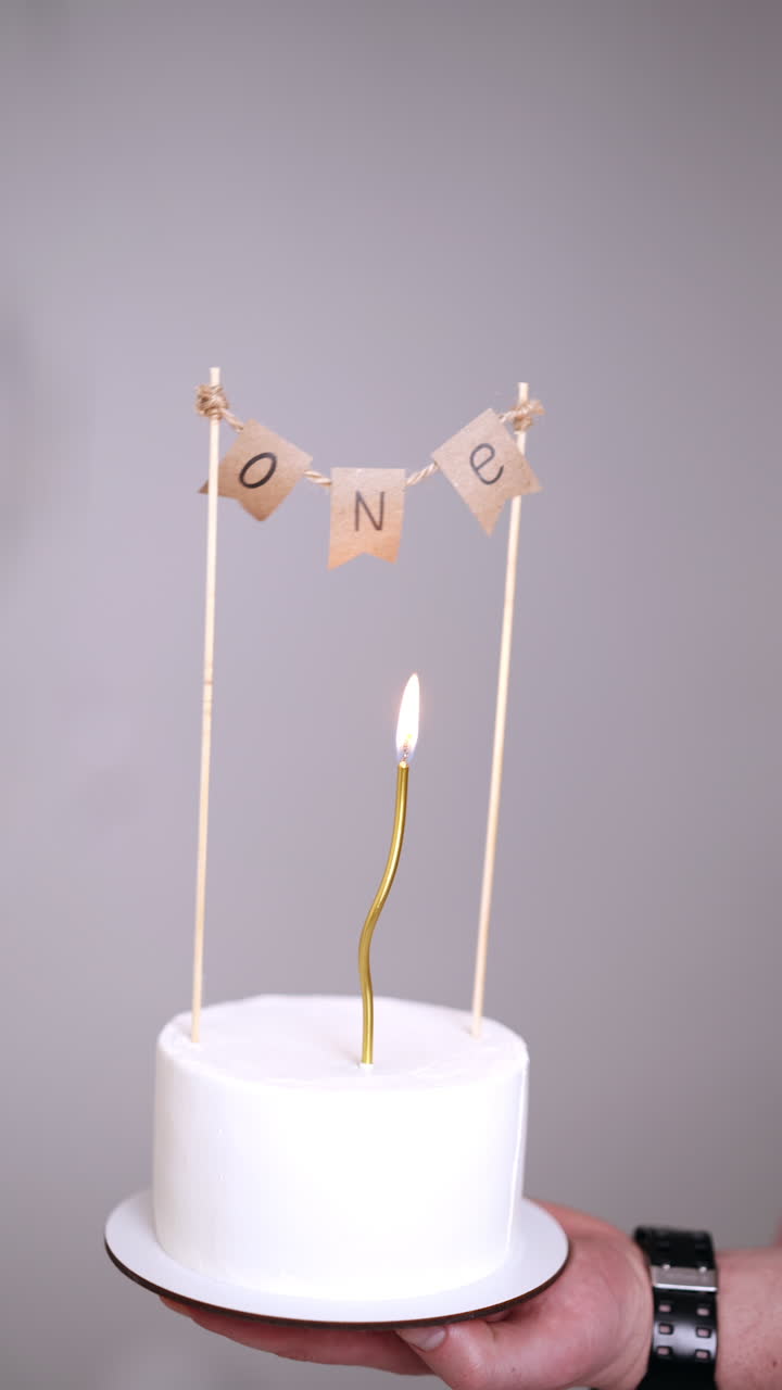 Male hand holding a white cake with a sign of one year celebration. A candle burning on the dessert for first baby birthday. White backdrop. Vertical video.