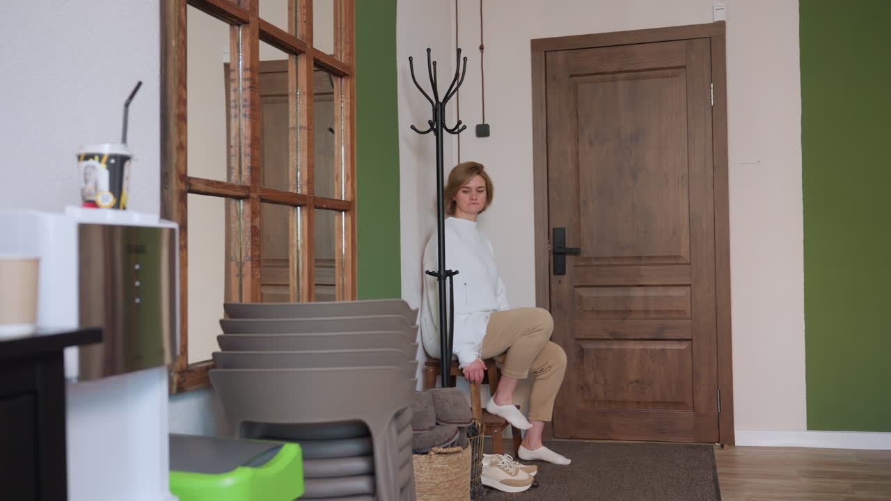 Female visitor stands from chair after removing canvas shoes and reaches for crocs in calm interior space with wooden door, coat rack, mirrors, and cozy seating area with stacked chairs