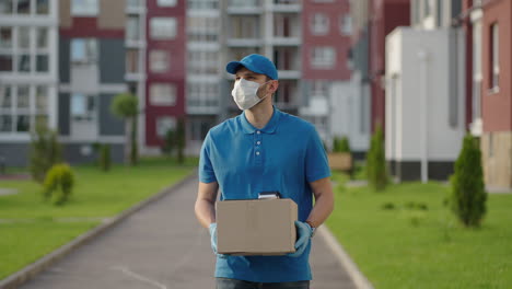 the male deliveryman in a cap and a protective mask and gloves goes with a box in his hands and carries a parcel to the customer. delivering online home orders.