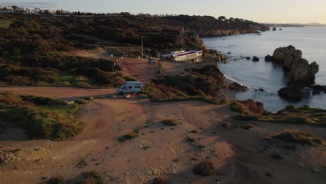 Aerial-view-orbiting-campervan-parked-at-Praia-dos-Arrifes-glowing-sunlit-sunrise-Portugal-shoreline