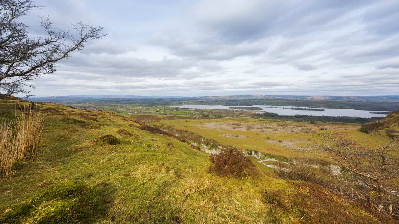 Panorama-timelapse af landligt landbrugslandskab med træer i forgrunden, græsmark, bakker og sø i det fjerne på en skyet dag, set fra Carrowkeel i Sligo County i Irland.