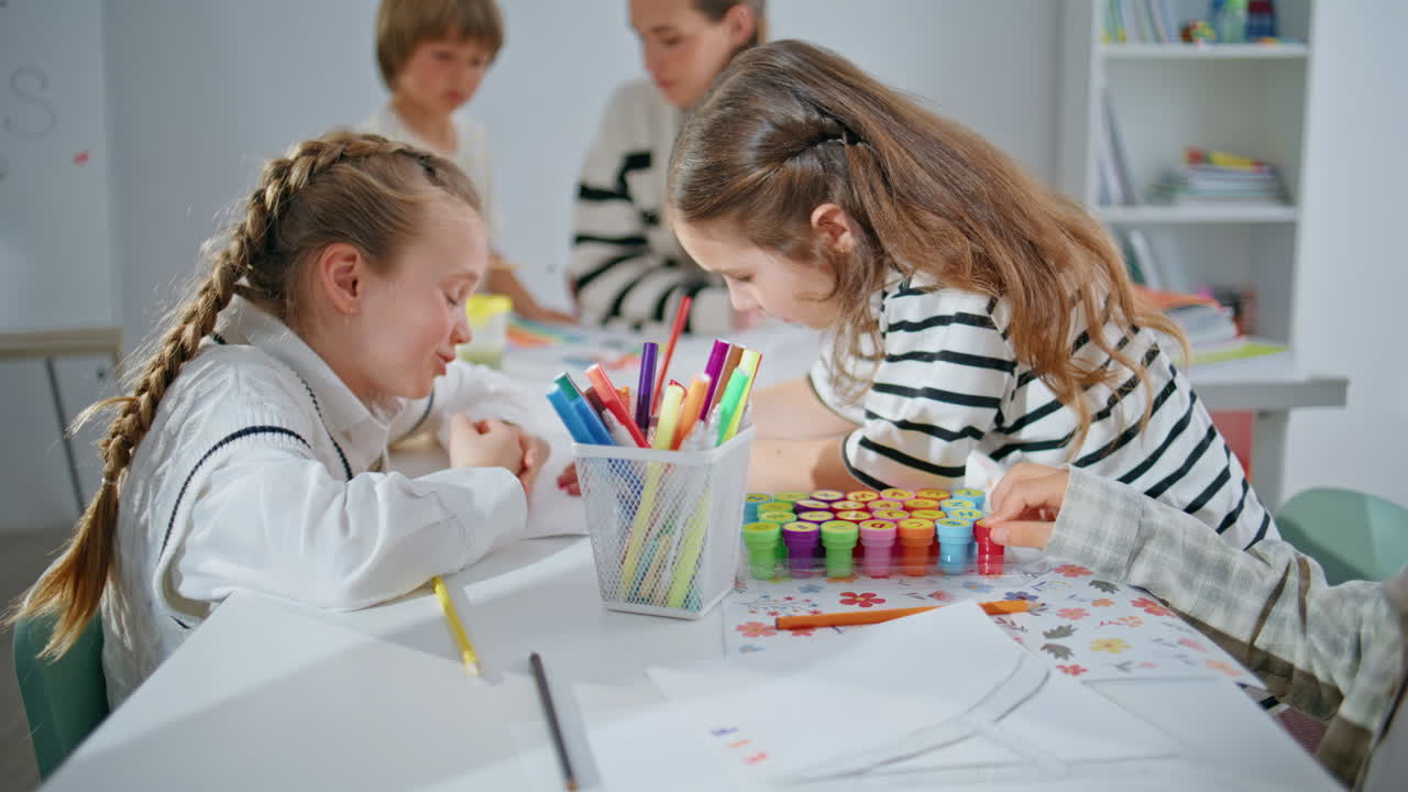Two girls drawing pictures at big school table closeup. Inspired kids talking