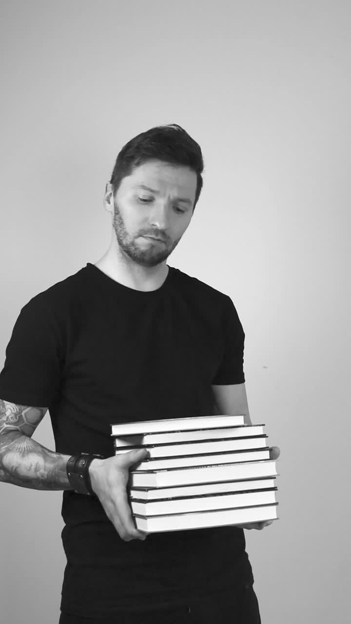 Grayscale footage of a tattooed short haired and bearded man holding and throwing a pile of hardcover or hardcopy books in his hand and arms in slow motion and acts surprised while watching at them.