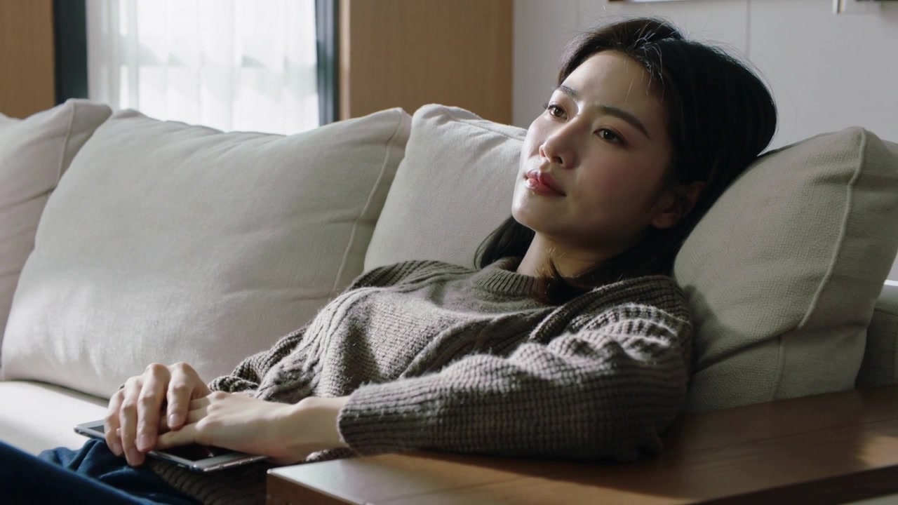 日差しが照らす中、茶色のセーターを着た女性がソファの上で頭を傾け、右を見つめながら携帯電話を持ち、物思いにふけっている。