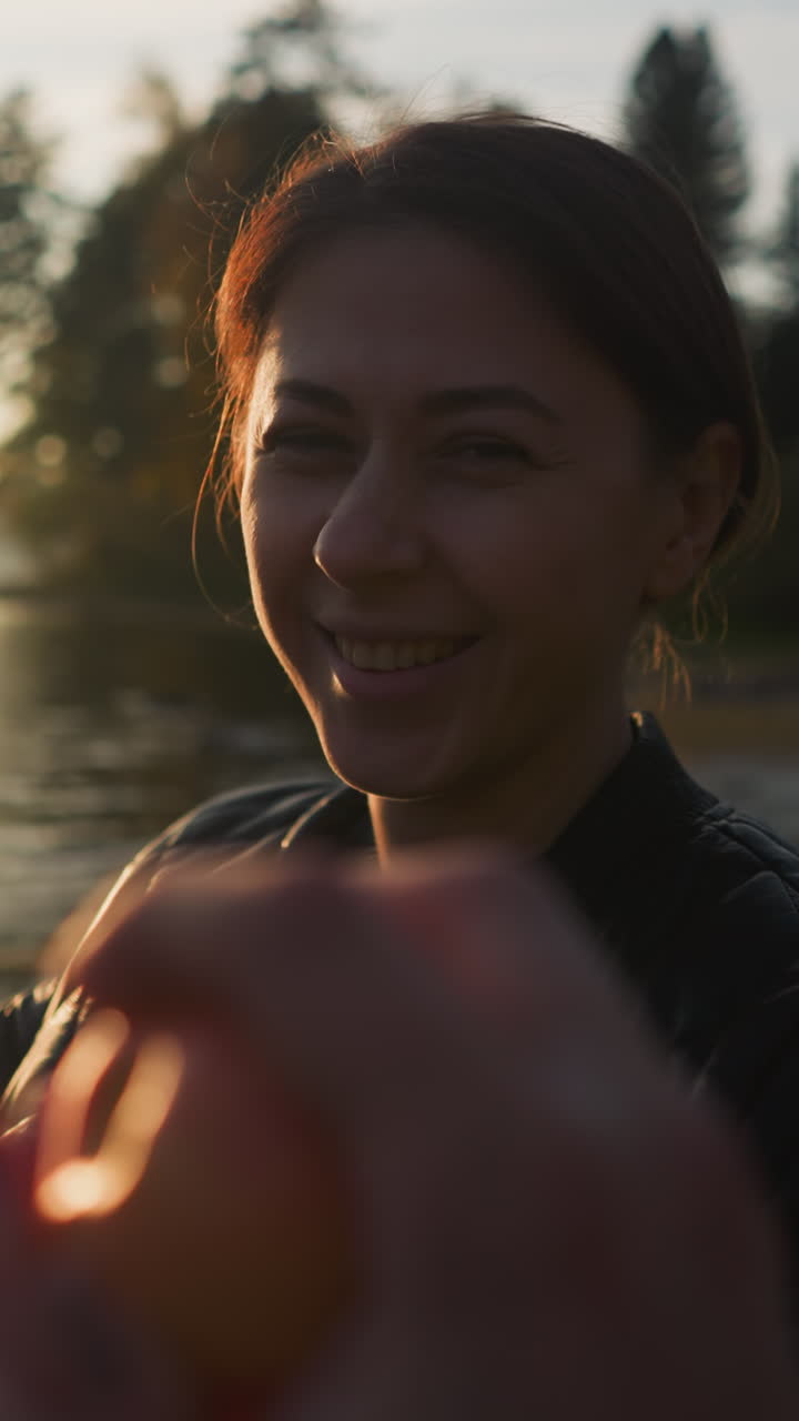 Lady gives apple to boyfriend on riverbank. Young woman offers fruit to partner silhouette resting near lake. Couple enjoys romantic atmosphere at sunset