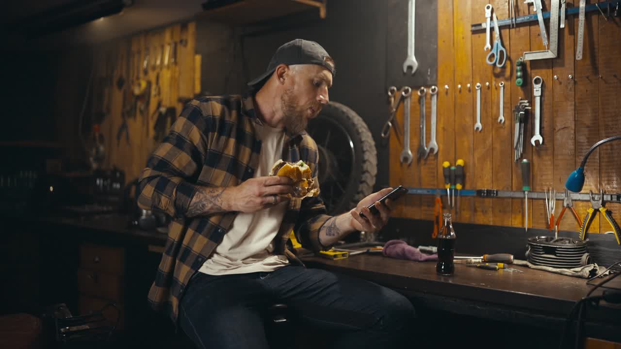 un tipo rubio con barba en una camisa a cuadros come una hamburguesa ve un video y bebe refrescos en su estudio de taller