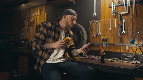 A-blond-guy-with-a-beard-in-a-plaid-shirt-eats-a-burger-watches-a-video-and-drinks-soda-in-his-workshop-studio