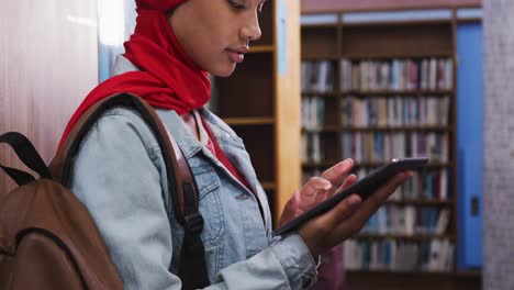 una estudiante asiática con un hijab rojo estudiando en una biblioteca y usando una tableta
