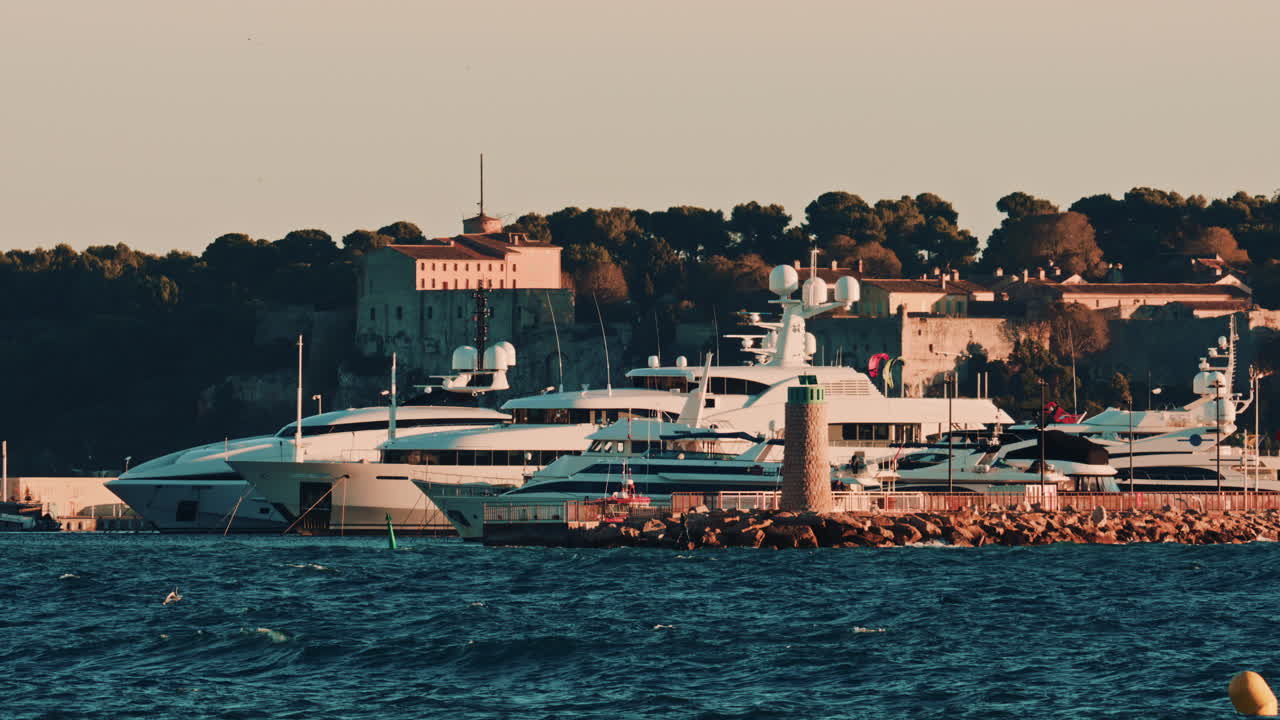 Luxury yachts docked near a stone lighthouse, with a historic building and trees in the background, port of Cannes, France