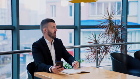 businessman in suit sitting at desk holding a phone in one hand and pen in another. man looks aside and talk strictly to someone.